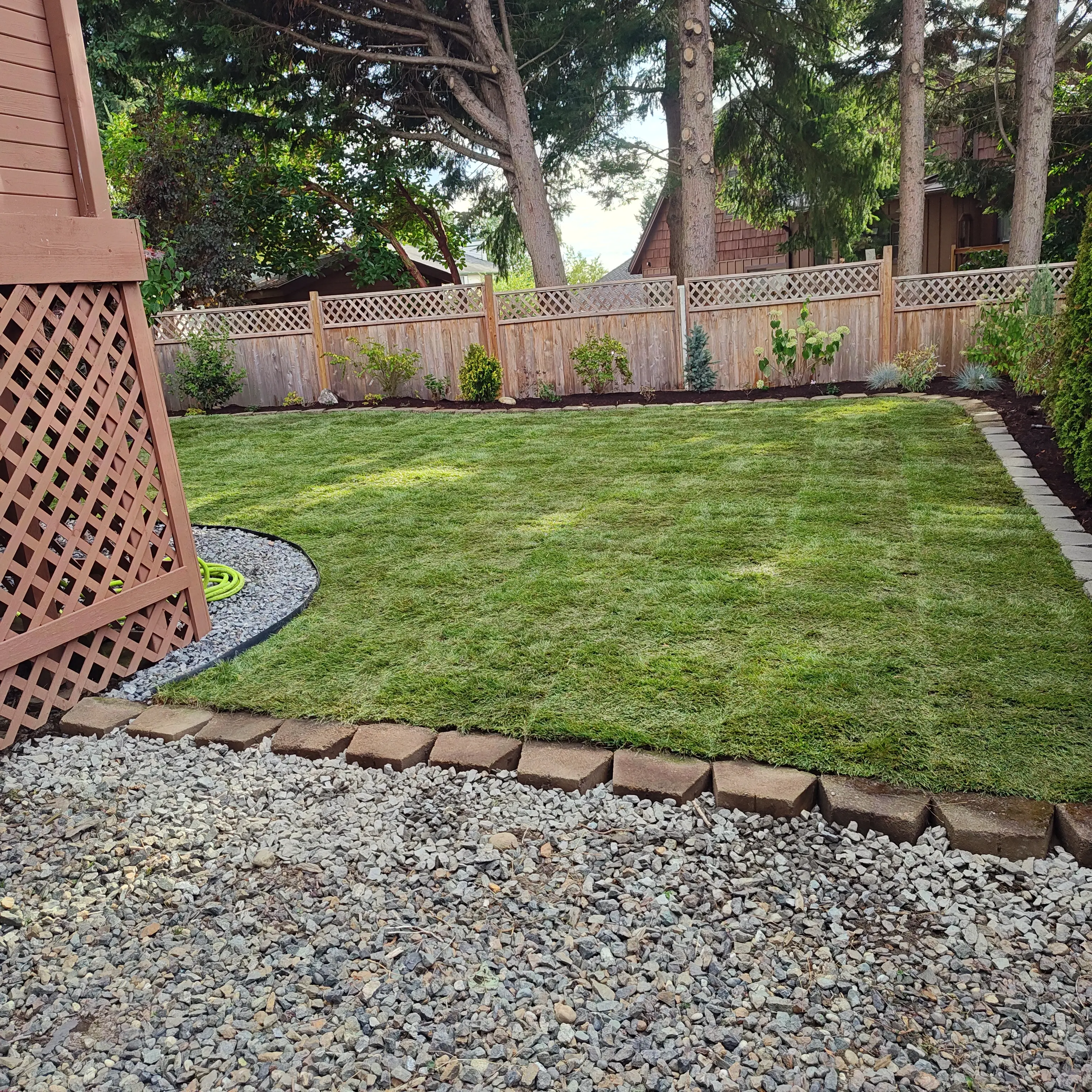 Beautiful backyard with fresh green lawn, wooden lattice fence with decorative top, landscaping along fence with shrubs and plants, stone edging, and gravel pathway.