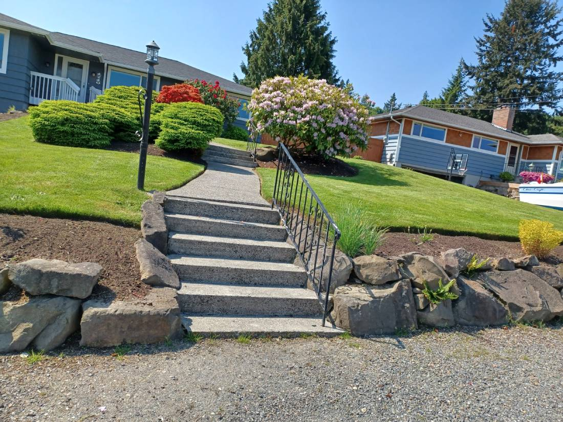 Concrete steps with metal railing, bordered by rock walls, mulch beds, and beautifully trimmed rounded shrubs with flowering plants.